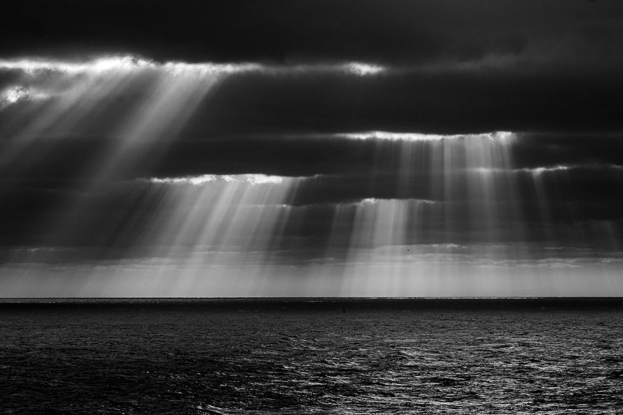 "Rébellion Lumineuse" est une photographie en noir et blanc saisissante issue de la série "Keshiki", capturant un moment unique où le soleil perce les nuages sombres au-dessus de la Manche, révélant une scène dramatique de lumière contre ténèbres.