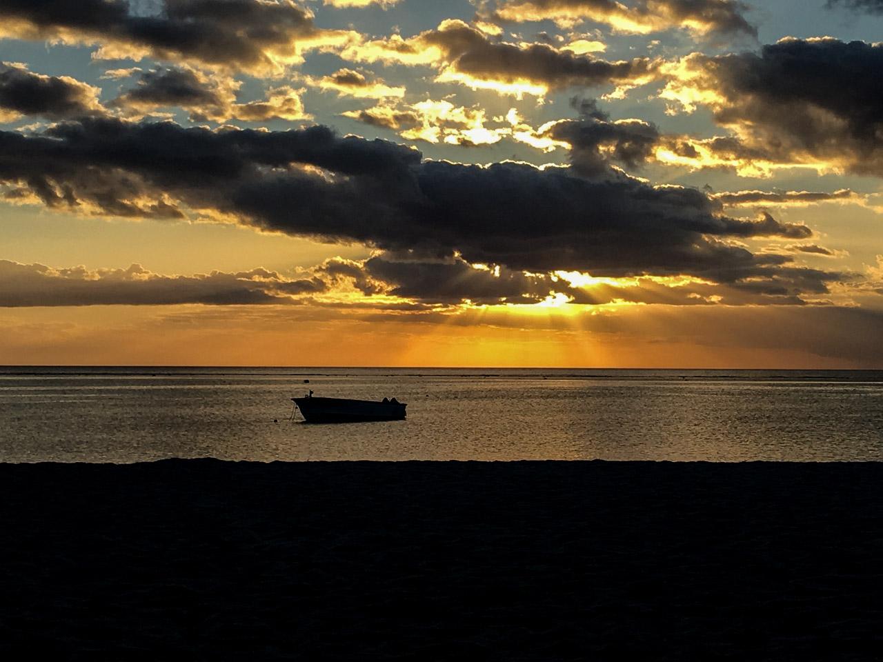 Lueur de l'Absence capture un coucher de soleil doré à l'île Maurice. La mer calme, une embarcation solitaire, et un ciel dramatique créent une scène nostalgique et sereine.