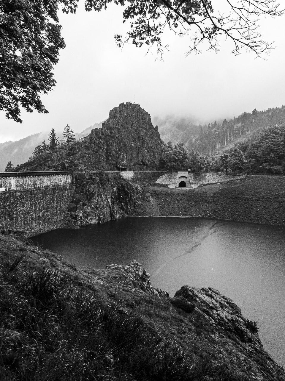 Sombre Résonance capture la beauté mélancolique du Gouffre d’Enfer sous un jour de pluie. Cette photo en noir et blanc dévoile un paysage où le barrage imposant se fond dans les roches naturelles, tandis que des sapins majestueux émergent du brouillard environnant.