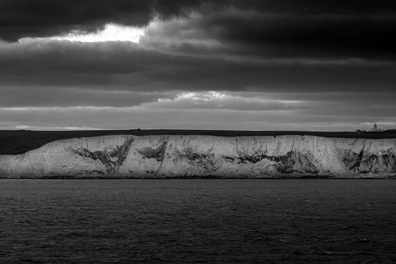 Wink White est une photographie en noir et blanc des falaises de Douvres, montrant un phare éclairé se dressant au-dessus d'une mer calme et d'un ciel nuageux.