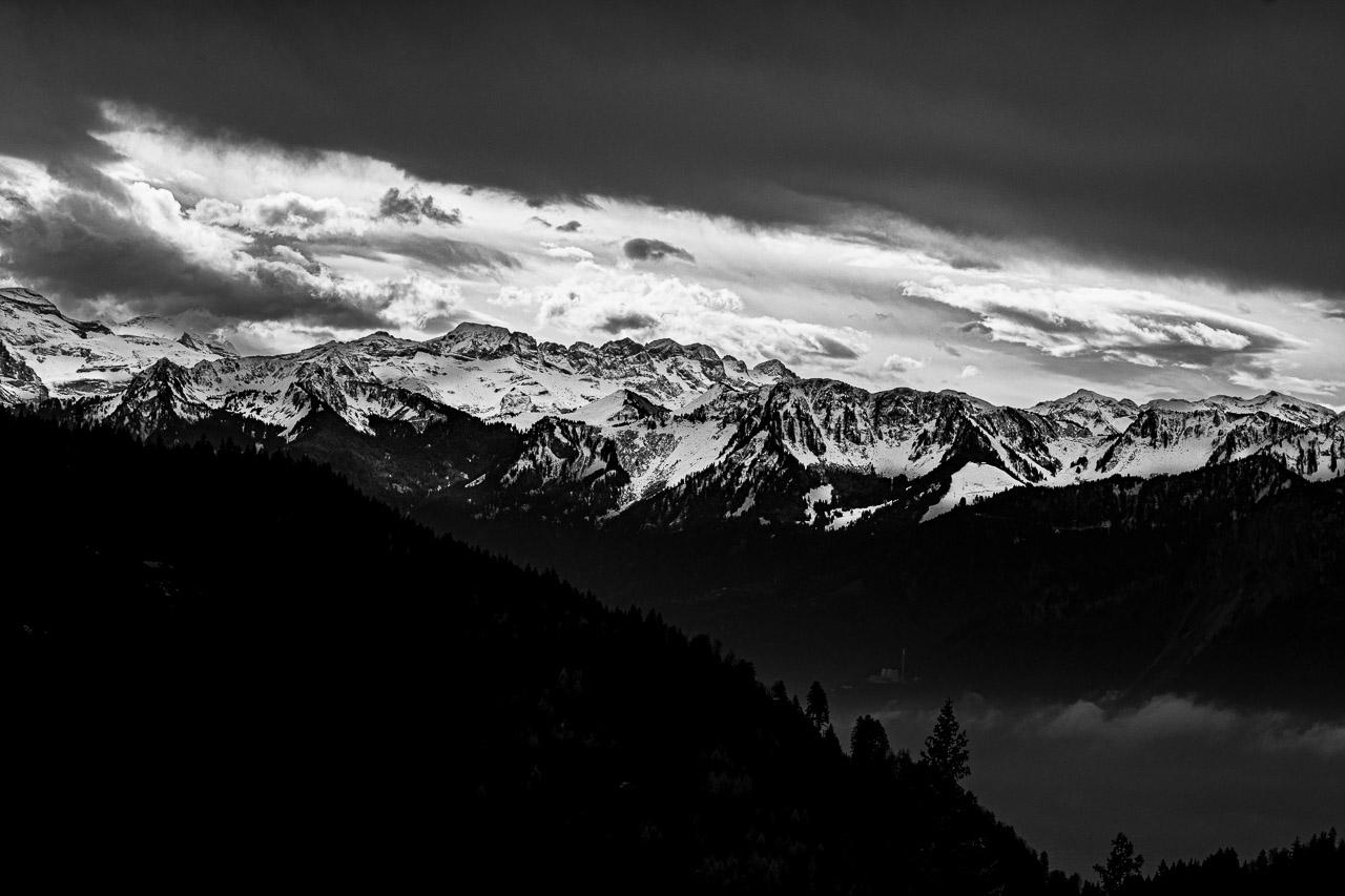 Entre Ombre et Murmure" capture la majesté des Alpes suisses en noir et blanc, révélant un paysage en trois bandes distinctes : une colline boisée en ombres chinoises, une montagne illuminée alternant neige et roche, et un ciel nuageux aux contrastes saisissants.